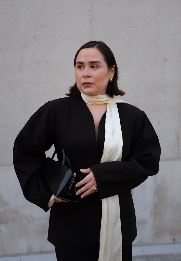 Blazer nero oversize con maniche a sbuffo, sciarpa crema annodata attorno al collo, borsa nera con accenti in argento, texture lisce, design minimalista.