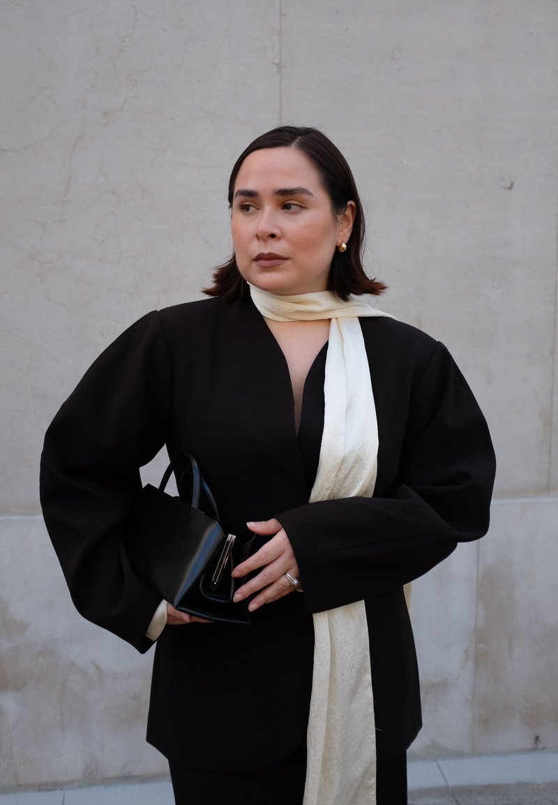 Blazer nero oversize con maniche a palloncino, sciarpa crema legata attorno al collo, borsa nera con dettagli in argento, texture morbide, design minimalista.