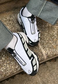 Bijele slip-on tenisice s crnim akcentima, sa zatvaranjem na patentni zatvarač i perforiranim dijelovima. Izrađene od izdržljivog materijala, mat površine i s podstavljenim đonom.