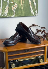 Um par de mocassins de couro castanho polido repousa numa consola de rádio vintage VAGNER de madeira, com flores secas decorativas nas proximidades.