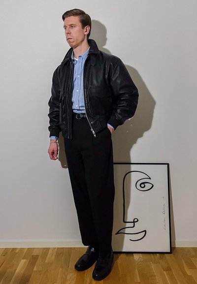 Chaqueta bomber de cuero negro sobre una camisa de rayas azul claro, pantalones negros y zapatos negros. Cerca hay un dibujo abstracto de un rostro enmarcado.
