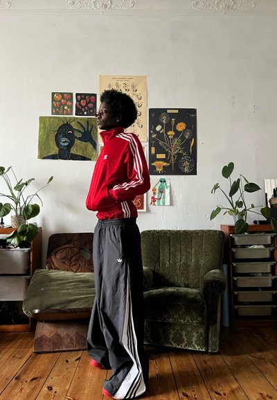 Chaqueta deportiva roja con cremallera y rayas blancas, combinada con pantalones de chándal negros holgados que presentan rayas y un logo similares. Ambiente interior con plantas.