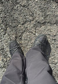 Personne portant un pantalon noir et des mocassins noirs debout sur un sol en asphalte rugueux.