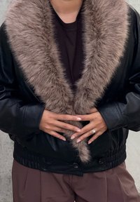 Casaco de couro preto com uma gola larga de pelo falso castanho. Apresenta punhos canelados e uma cintura ajustada. Mãos posicionadas à frente, usando um anel.