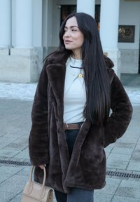 Manteau en fausse fourrure marron avec un large revers, porté sur un haut blanc. Il est accompagné d'une ceinture marron et d'un sac à main beige, sur fond de pierre.