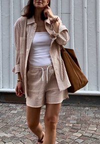 Nő bézs és fehér csíkos sortot és illő, oversized inget visel, fehér topot, barna válltáskát, és macskaköves úton sétál.