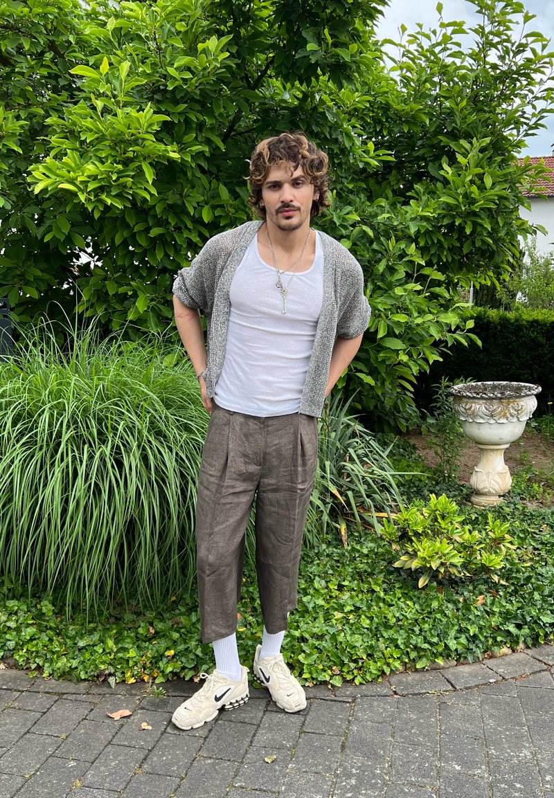 Jeune homme aux cheveux bouclés, debout sur un chemin de pierre, portant un cardigan gris, une chemise blanche, un pantalon brun ample, des chaussettes blanches et des baskets beiges, avec de la verdure en arrière-plan.