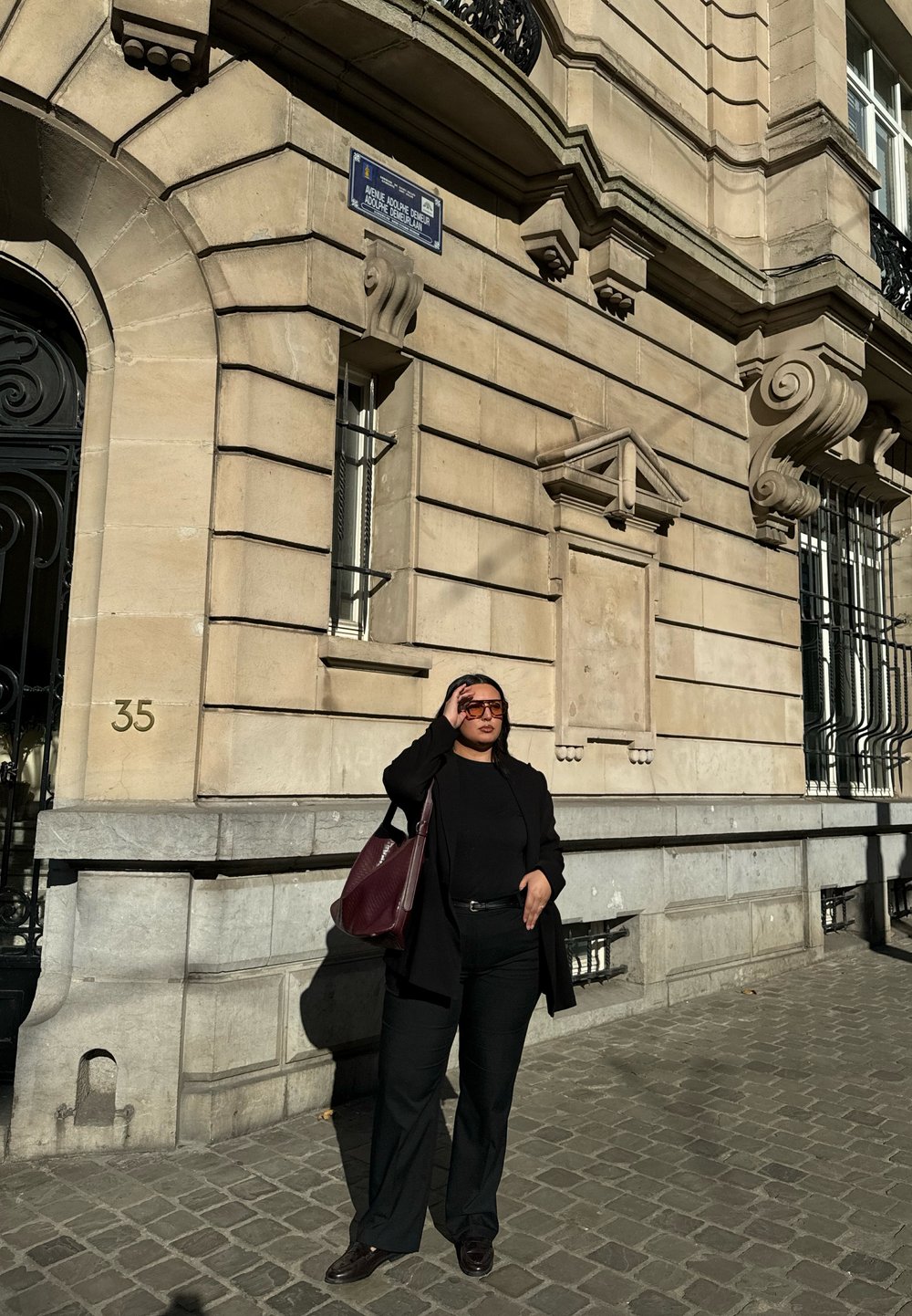 Bâtiment avec une façade en pierre de couleur claire, détails ornés et une porte en métal ; une personne en tenue noire et un sac bordeaux se tient contre cela.