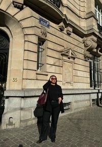 Bâtiment avec une façade en pierre de couleur claire, détails ornés et une porte en métal ; une personne en tenue noire et un sac bordeaux se tient contre cela.