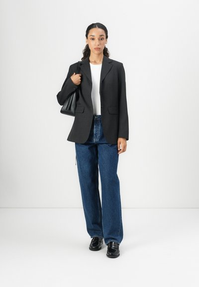 Blazer negro con un diseño estructurado, combinado con una blusa blanca y jeans de mezclilla azul de pierna ancha. El conjunto se completa con zapatos negros y un bolso.