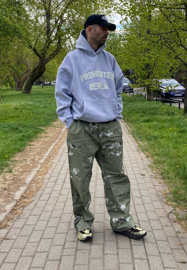 Mies, jolla on harmaa "Prohibited Berlin" -huppari, maastokuvioiset cargohousut, musta lippalakki ja tennarit, seisoo puistopolulla vihreiden puiden ympäröimänä.