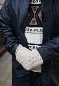 Czarna wodoodporna kurtka, dziergany sweter z geometrycznymi wzorami w kolorach brązowym i zielonym oraz kremowe mitenki z warkoczowym splotem. Ciepłe tekstury i warstwowe projekty.