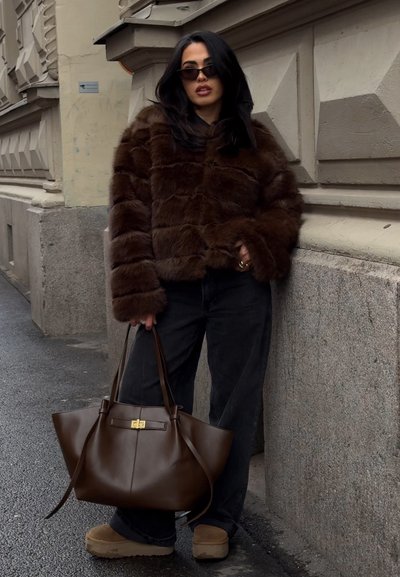 Mujer vestida con una chaqueta de piel marrón oscuro y jeans negros sostiene un gran bolso de cuero marrón, de pie en la acera de la ciudad junto a una pared texturizada.