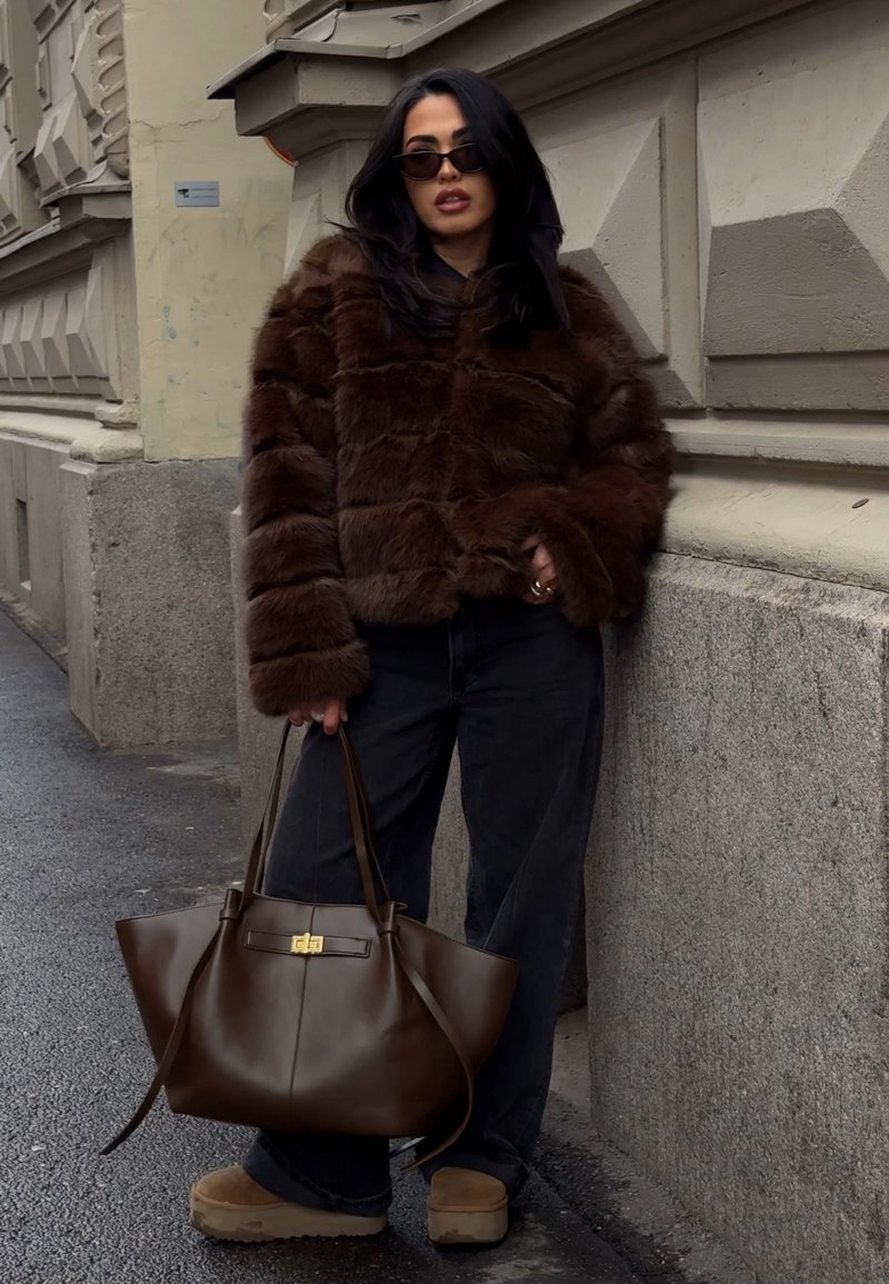 Woman wearing dark brown fur jacket and black jeans holds large brown leather handbag, standing on city sidewalk by textured wall.