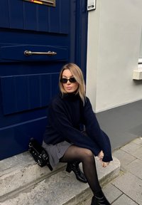 Tengerészkék pulóver, szürke szoknya, fekete vastag harisnya és bokacsizma. Fekete táska fém részletekkel a kő lépcsőkön áll egy kék ajtó mellett.