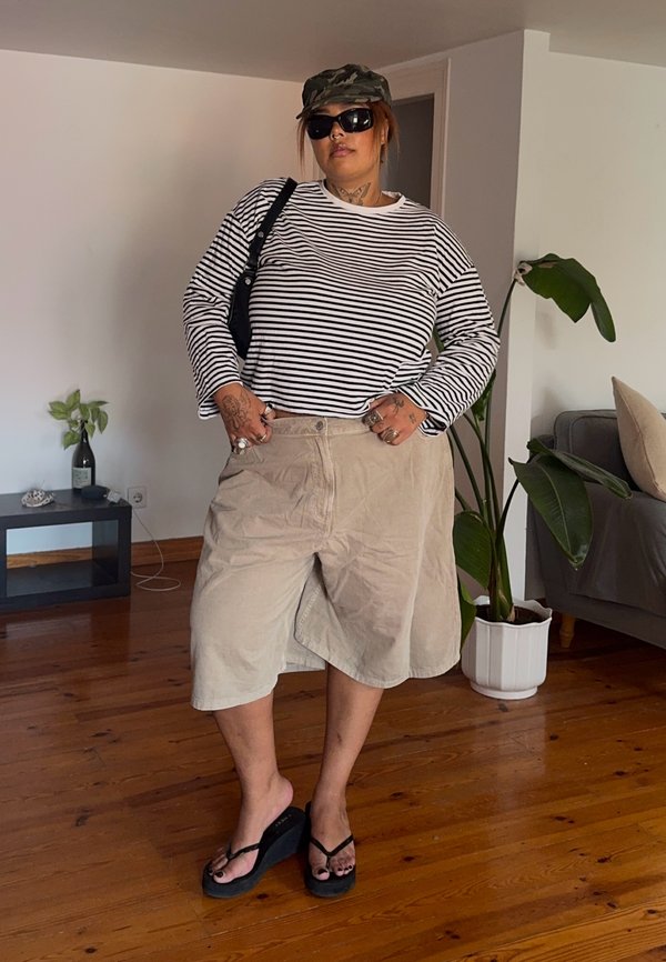 Person iført en langærmet, stribet skjorte, beige vide shorts, sorte solbriller, camouflage kasket og sorte kilehæls-sandaler, stående indendørs.