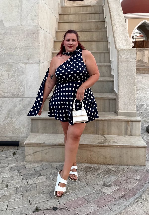 Femme en robe bleu marine à pois et sandales blanches, debout sur des marches en pierre, tenant un petit sac à main blanc, avec des bâtiments publics en arrière-plan.