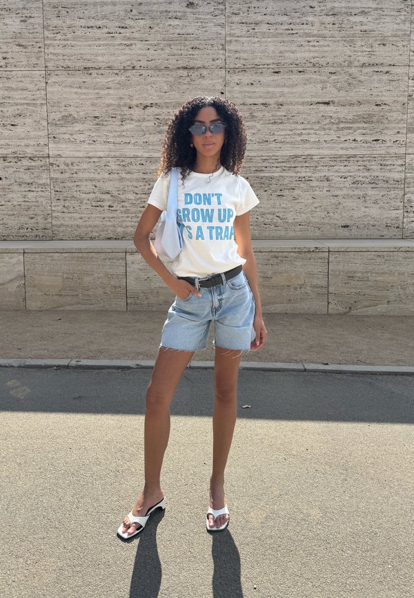 Vrouw met krullend haar die een zonnebril draagt, wit t-shirt met tekst, spijkershorts, witte sandalen en een schoudertas die buiten bij een stenen muur staat.