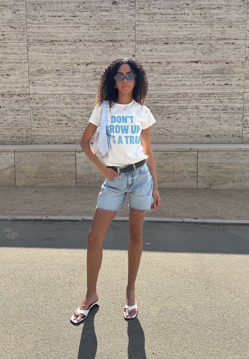Femme aux cheveux bouclés portant des lunettes de soleil, un t-shirt blanc avec un texte, un short en denim, des sandales blanches et un sac à bandoulière, debout dehors près d'un mur en pierre.