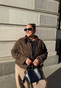 Jaqueta de camurça castanha com fecho de zip, calças largas bege e uma mala preta. Os acessórios incluem anéis e óculos de sol. Fundo urbano iluminado pelo sol.