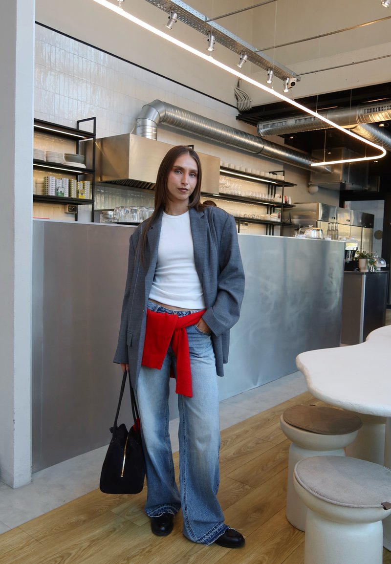 Jeune femme en blazer gris, chemise blanche et pull rouge noué à la taille, debout tenant un sac noir dans un café moderne avec une décoration métallique et en bois.