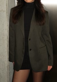 Tume roheline täpselt sobiv bleiser kahe nööbiga, ees taskud ja sile kangas, kantud musta kõrge kaelusega pluusi ja lühikese seelikuga.