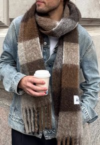 Écharpe à carreaux marron et gris en matériau doux, avec des franges aux extrémités et une étiquette de logo. Portée avec une veste en jean et tenant une tasse de café.
