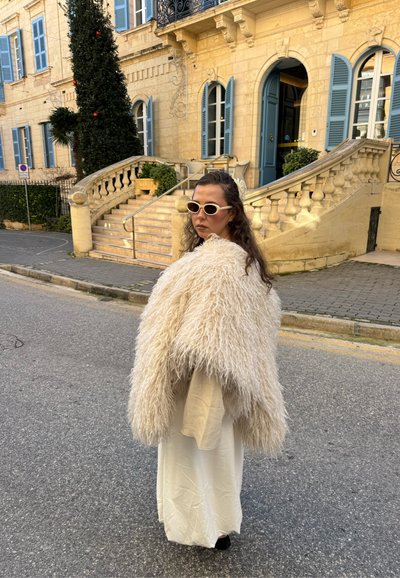Mujer con abrigo de piel blanco, vestido beige y gafas de sol, de pie en la calle cerca de una escalera de piedra y un edificio amarillo con contraventanas azules.