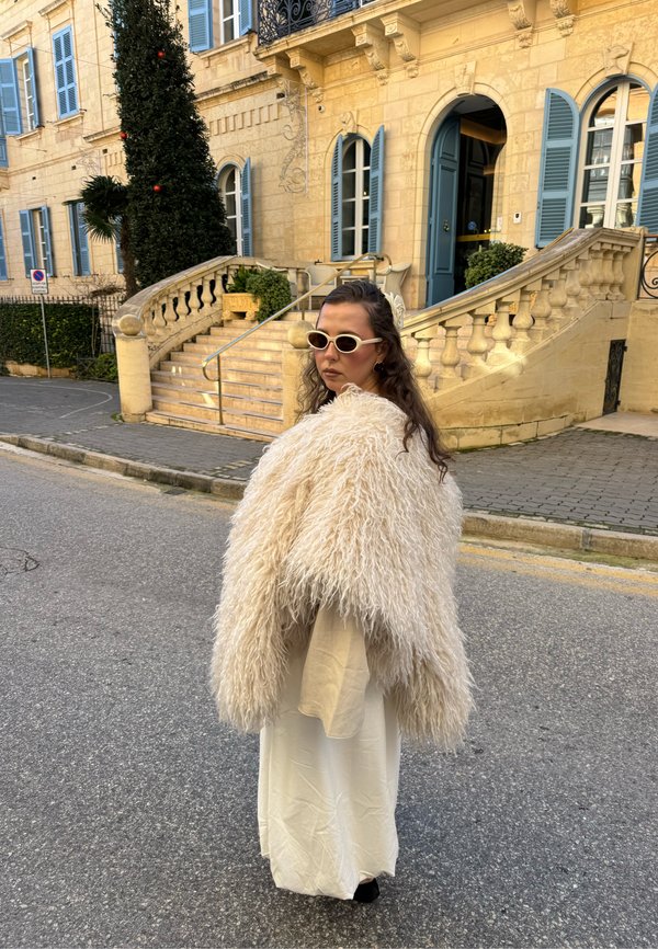 Donna con cappotto di pelliccia bianca, vestito beige e occhiali da sole in piedi sulla strada vicino a una scala di pietra e a un edificio giallo con persiane blu.