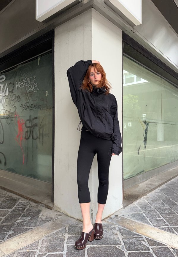 Femme aux cheveux rouges portant une veste noire, des leggings courts et des sabots à talons, appuyée contre un coin d'un bâtiment urbain avec des graffitis sur la fenêtre.
