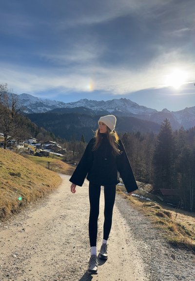 Mujer con chaqueta negra, leggings y gorro blanco caminando por un sendero de montaña con picos nevados y cielo iluminado por el sol al fondo.
