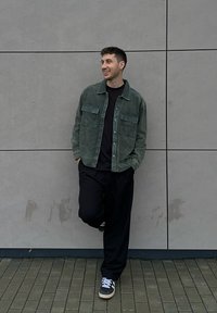 Veste-chemise en velours côtelé vert avec deux poches poitrine sur un t-shirt noir, associée à un pantalon noir ample et des baskets grises sur un sol en briques texturé.