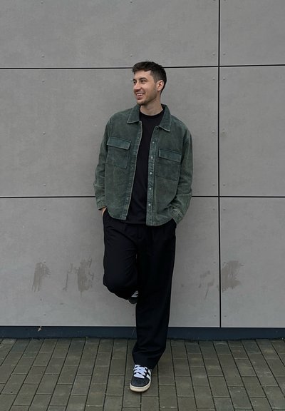 Sobrecamisa de pana verde con dos bolsillos en el pecho sobre una camiseta negra, combinada con pantalones negros holgados y zapatillas grises sobre un suelo de ladrillo texturizado.