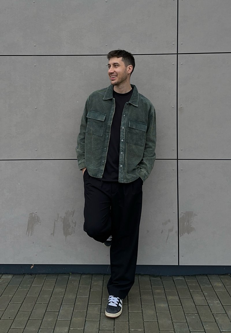 Veste-chemise en velours côtelé vert avec deux poches poitrine sur un t-shirt noir, associée à un pantalon noir ample et des baskets grises sur un sol en briques texturé.