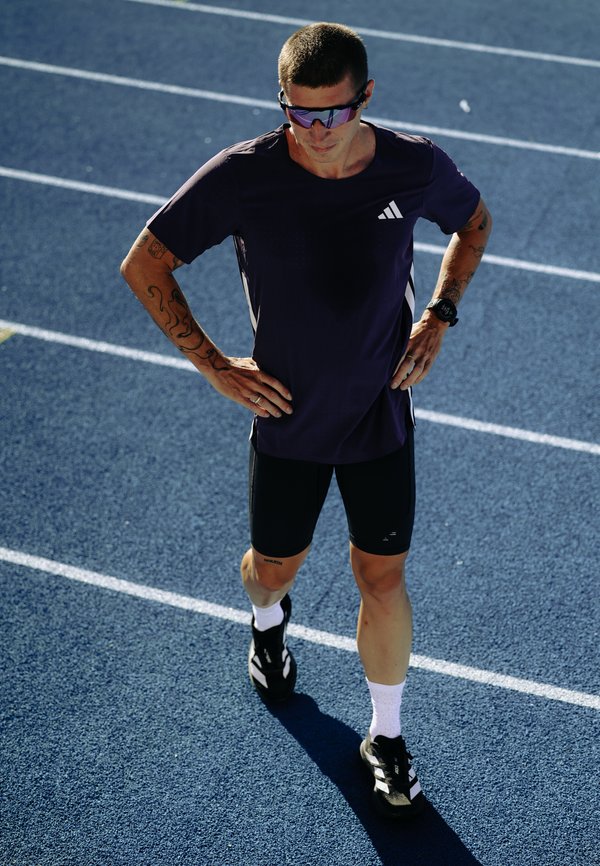 Mandlig atlet iført solbriller og sportstøj, stående på en blå løbebane med hænderne på hofterne, forbereder sig til træning.