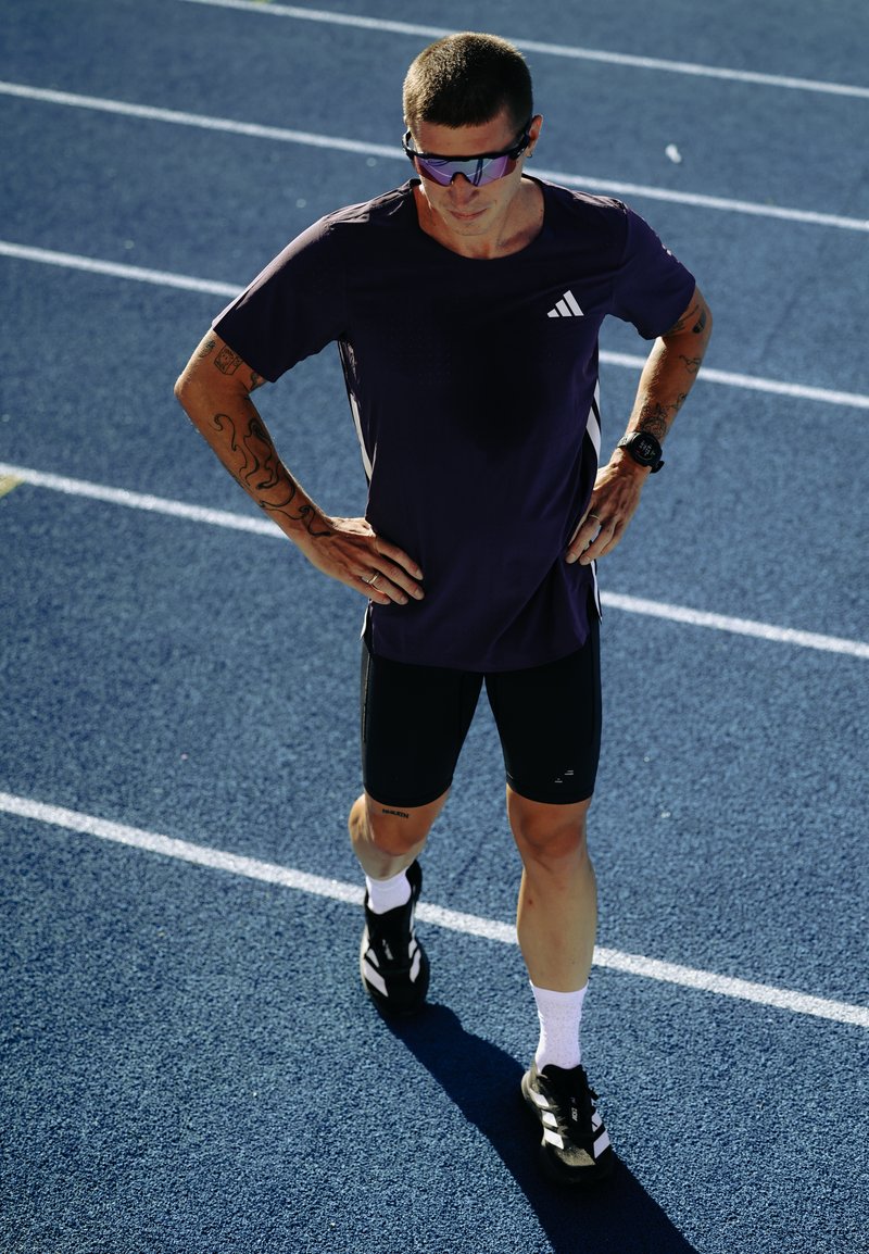 Mandlig atlet iført solbriller og sportstøj, stående på en blå løbebane med hænderne på hofterne, forbereder sig til træning.