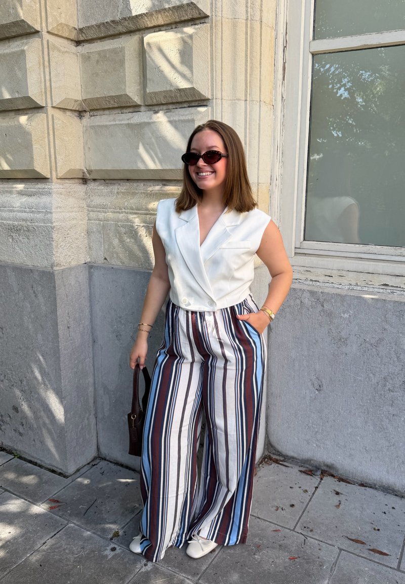 Femme en lunettes de soleil portant un blazer blanc sans manches et un pantalon large rayé, souriante près d'un bâtiment en pierre avec une fenêtre.