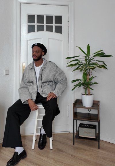 Chaqueta de denim gris con dos bolsillos frontales, camiseta blanca, pantalones negros holgados y zapatos negros. Planta en una maceta blanca al lado de un soporte de madera.