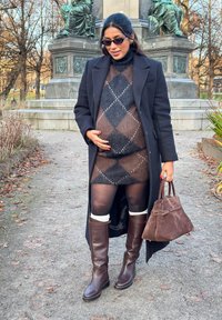 Brune knelange skinnstøvler, brunt mønstrete kjole, svart frakk; holder en brun semsket veske. Står på en grussti med trær.