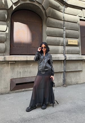 Chaqueta bomber negra sobre una falda larga negra semitransparente, combinada con zapatos negros de suela gruesa. El fondo presenta una pared de piedra texturizada y puertas cerradas.