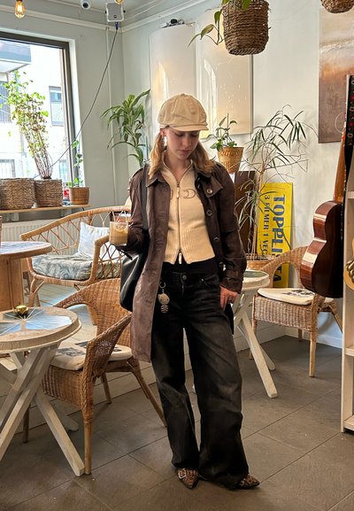 Abrigo de trench de cuero marrón, suéter cremoso con cremallera, jeans negros de pierna ancha y zapatos estampados. Sosteniendo una bebida, de pie en una cafetería con plantas.