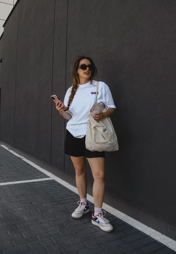 T-shirt bianco oversize con logo, pantaloni corti neri, borsa a spalla crema e sneakers multicolori. In piedi contro un muro scuro.