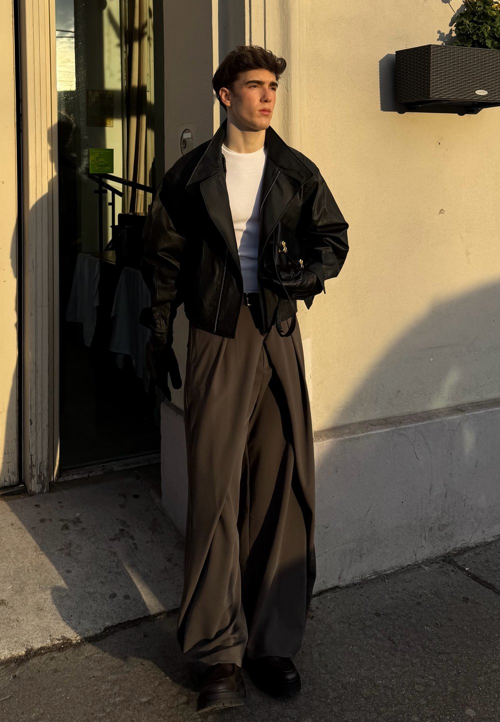 Veste courte en cuir noir portée sur un t-shirt blanc, associée à un pantalon ample gris. Des gants et des chaussures foncées complètent la tenue. Une lumière chaude projette des ombres.