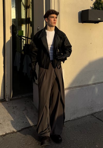 Chaqueta corta de cuero negro sobre camiseta blanca, combinada con pantalones sueltos grises. Guantes y zapatos oscuros completan el conjunto. La luz cálida proyecta sombras.