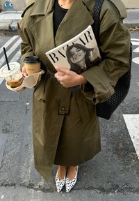 Pessoa com um casaco trench verde-azeitona a segurar a revista Harper's Bazaar e dois copos de café, vestindo sapatos brancos e pretos com manchas e carregando uma bolsa de vime.