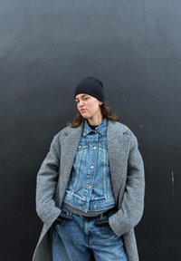 Personne portant un bonnet noir, un manteau gris par-dessus une veste en denim et un jean, debout les mains dans les poches contre un mur gris foncé.
