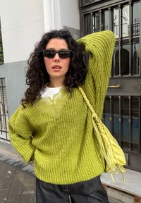 Femme aux cheveux foncés bouclés portant des lunettes de soleil noires, un pull épais vert citron, un jean foncé et un sac à main tressé assorti jaune-vert.