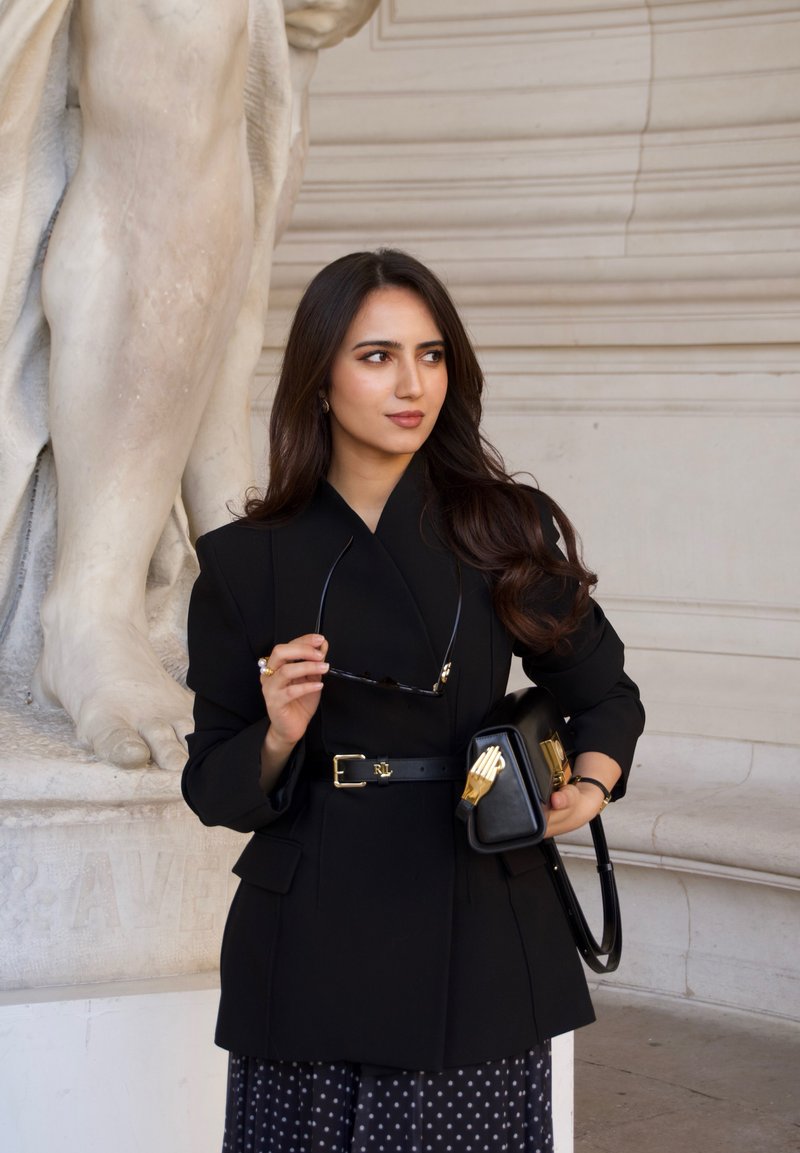 Femme en blazer noir et jupe à pois tenant des lunettes de soleil et un sac noir, debout à côté d'une grande statue en marbre blanc.