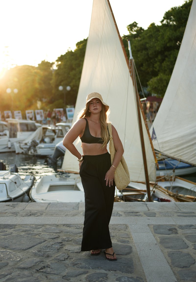 Top court noir, pantalon noir fluide, chapeau en paille à larges bords et sac en osier ; debout près des bateaux, avec un coucher de soleil en arrière-plan et des voiles en vue.