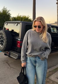 Jeune femme portant des lunettes de soleil, un pull gris et un jean, tenant un sac à main noir à côté d'un véhicule tout-terrain noir au coucher du soleil.